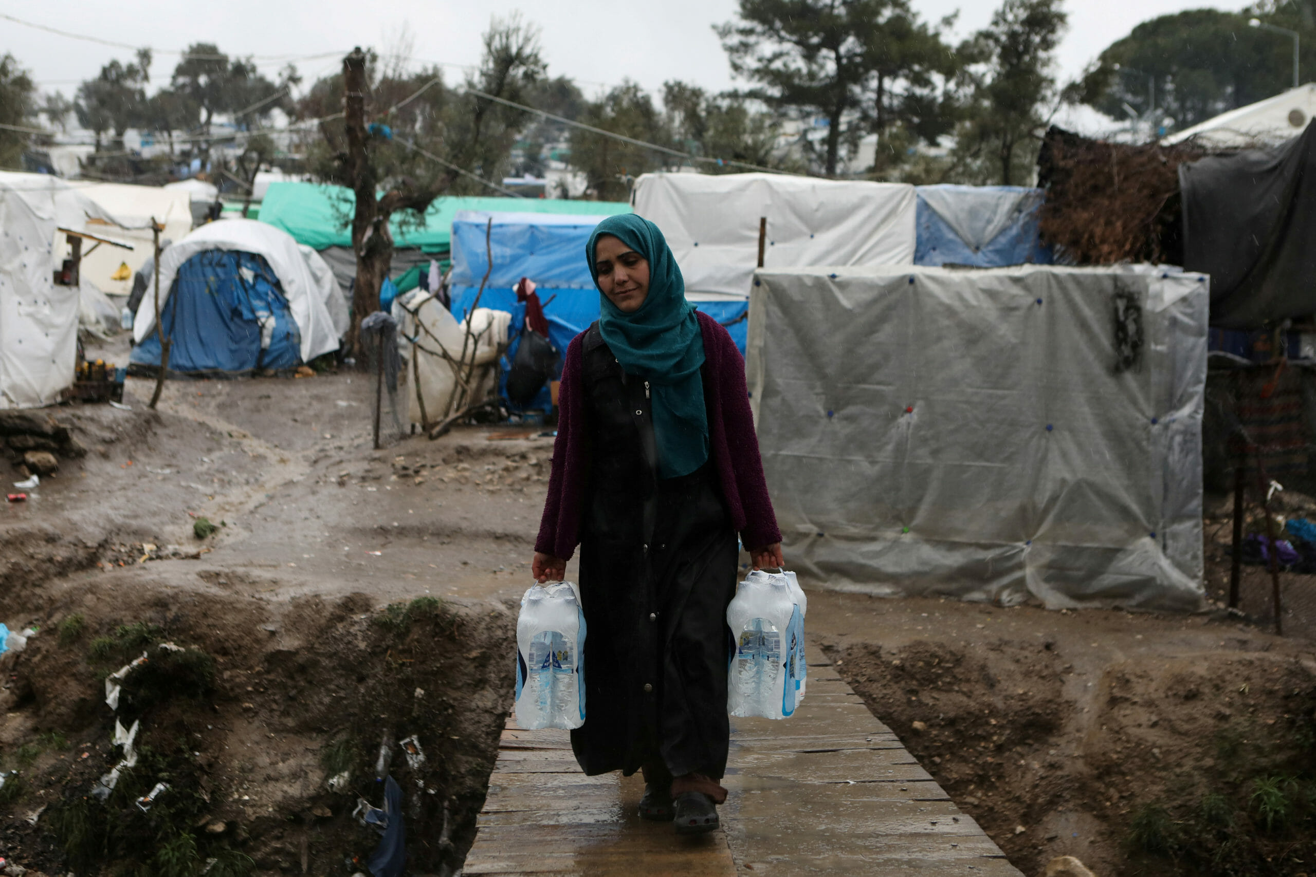 FILE PHOTO: A woman carries bottles of water during heavy rainfall at a temporary camp for refugees and migrants next to the Moria camp on the island of Lesbos, Greece, February 6, 2020. REUTERS