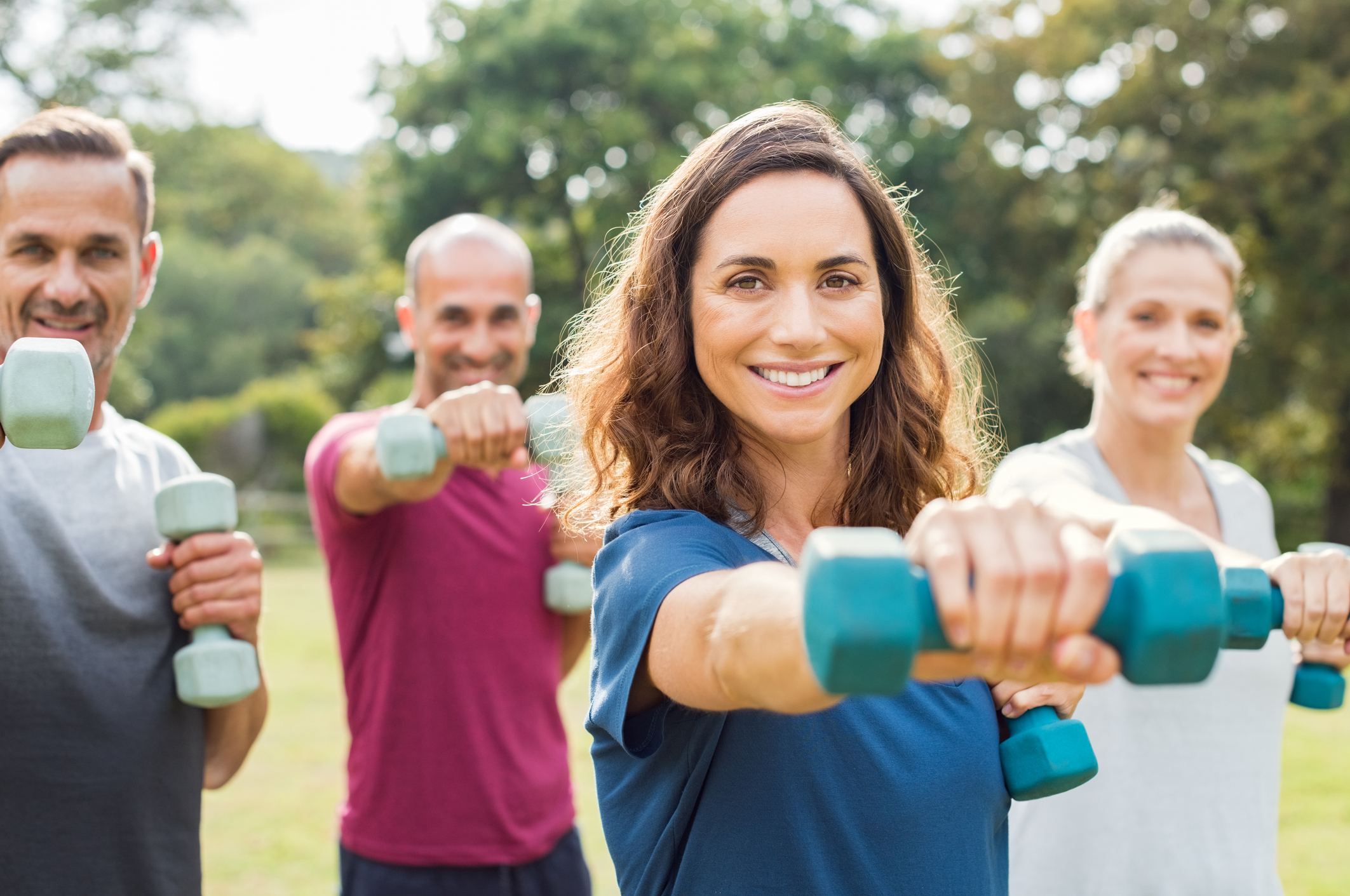 Mature people in training session of aerobics using dumbbells at park. Happy man and smiling woman practicing fitness together outdoor. Portrait of mature woman doing exercise with other people in background.