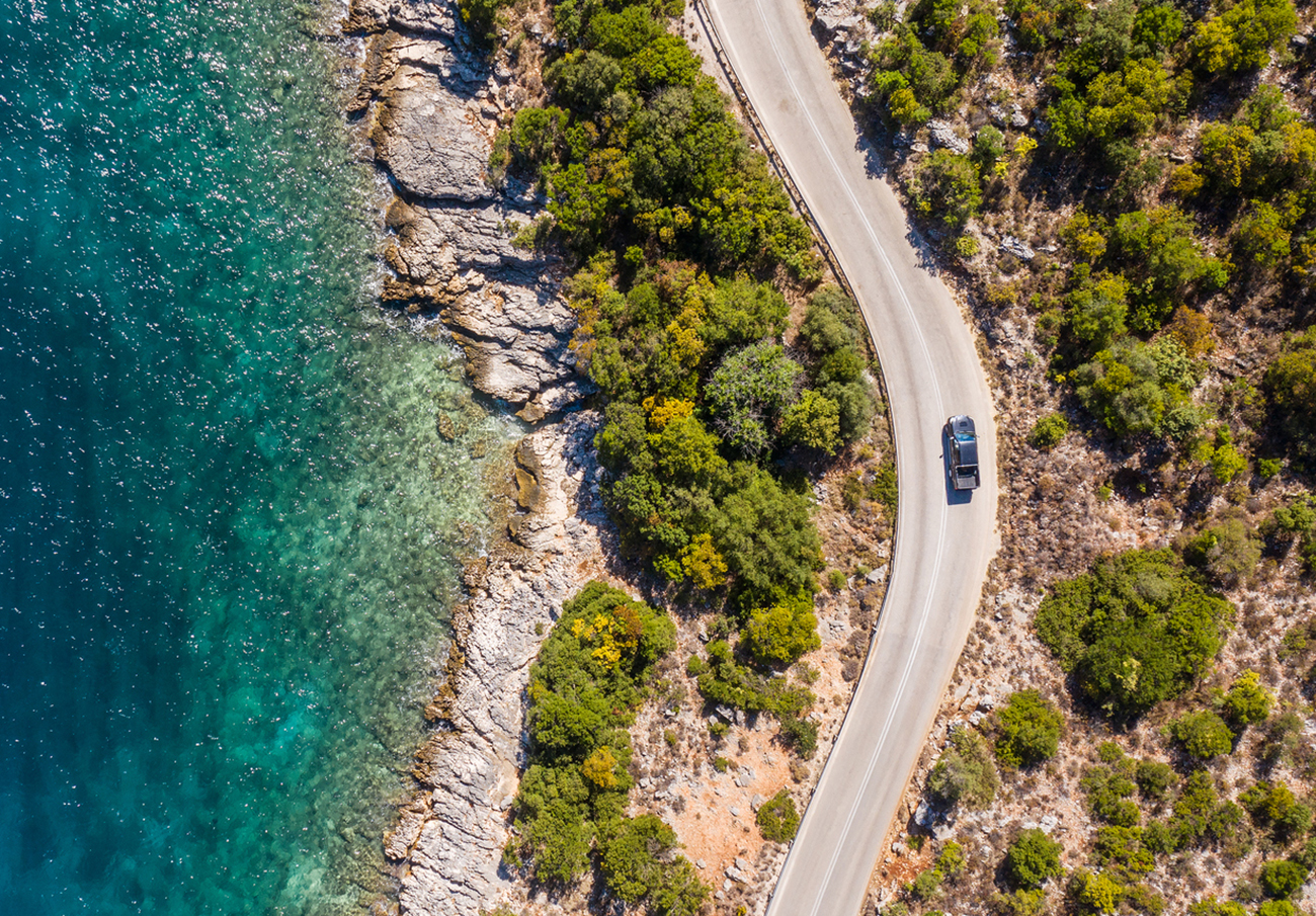 Aerial vertical shot of pickup truck moving by the curved road near sea tranquil waves on coast on Cephalonia Greek island. Transportation, traveling and nature concept.