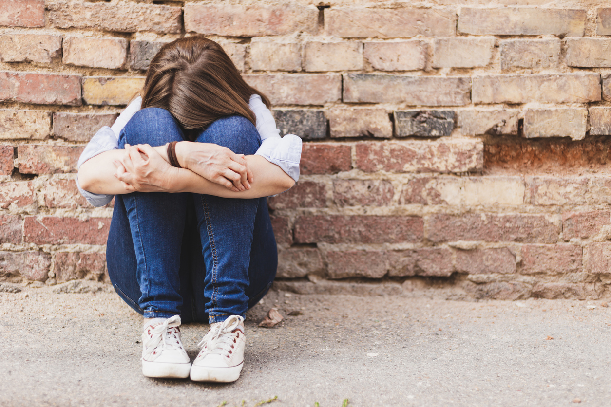 Scared millennial girl sitting alone in the city next to a brick wall - Casually dressed young woman wearing jeans hiding outdoors - Insecure female having panic attack