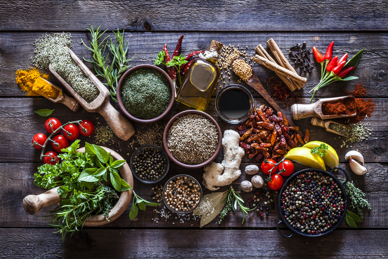 Top view of a rustic wood kitchen table filled with a large group of multi colored spices and herbs in bowls, wooden serving scoops or placed directly on the table. Spices and herb included are clove, turmeric, bay leaf, cinnamon, olive oil, curry powder, ginger, nutmeg, peppercorns, cinnamon, salt, chili pepper, basil, parsley, lemon, rosemary, garlic, onion and saffron. Low key DSRL studio photo taken with Canon EOS 5D Mk II and Canon EF 100mm f