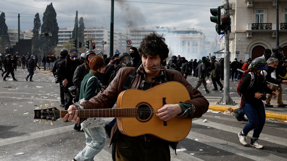 Μουσικός παίζει κιθάρα ενώ πίσω του εκτυλίσσονται επεισόδια