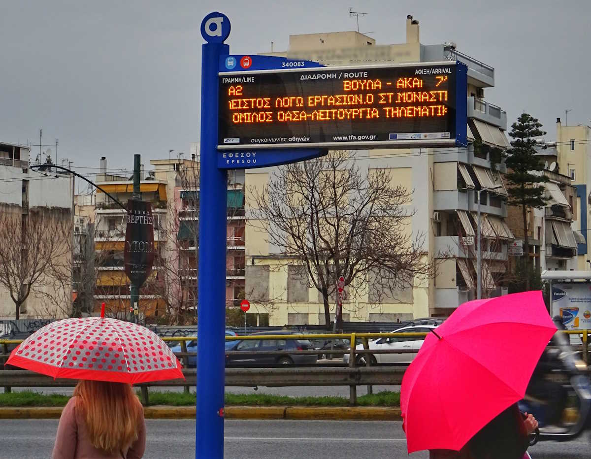 Αθηναίοι περιμένουν σε στάση λεωφορείου με την ηλεκτρονική ένδειξη να αναγράφει τον χρόνο άφιξης του μέσου μαζικής μεταφοράς