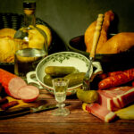 A stylized antique still life with meat, sausages, salami, lard and a glass of vodka