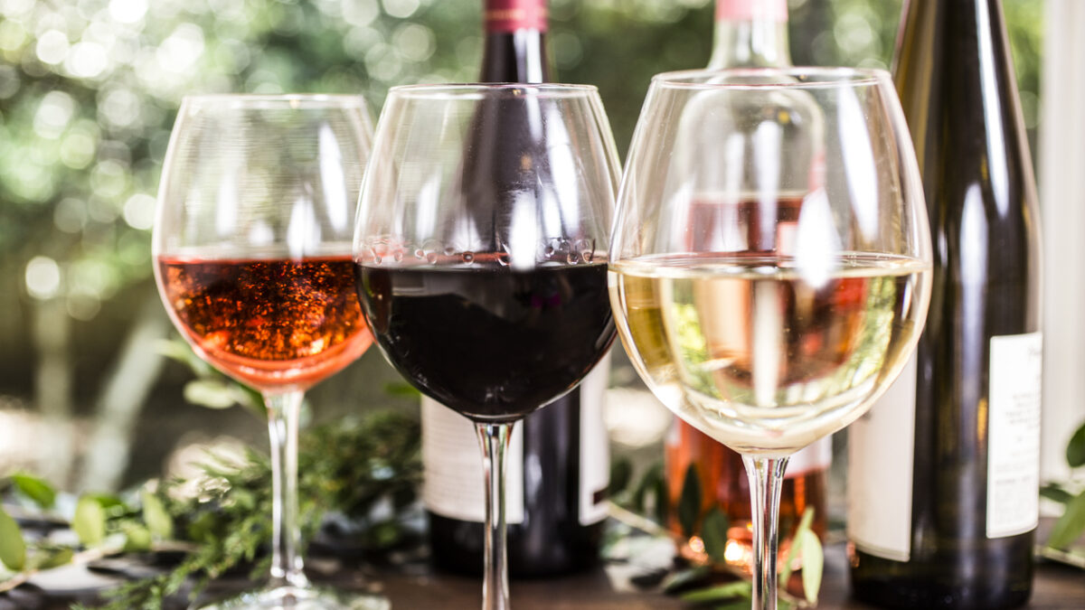 Wine tasting event. White, rose and red wines in glasses on outdoor patio table. Various wine bottles in background including white, red, and rose selections. Rustic wooden table with vineyard grounds in background. No people.