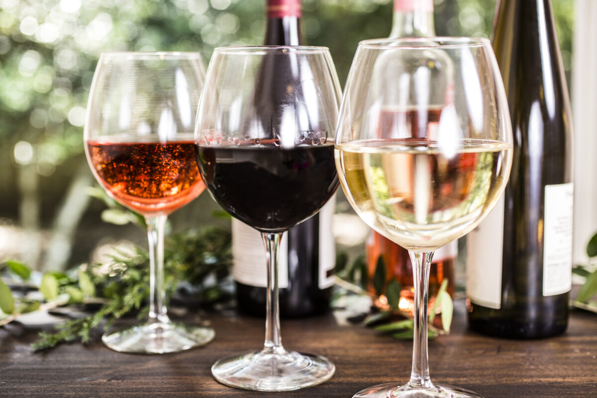 Wine tasting event. White, rose and red wines in glasses on outdoor patio table. Various wine bottles in background including white, red, and rose selections. Rustic wooden table with vineyard grounds in background. No people.