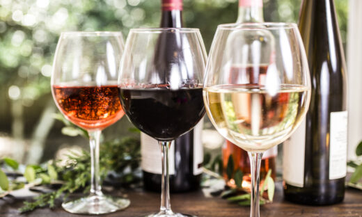 Wine tasting event. White, rose and red wines in glasses on outdoor patio table. Various wine bottles in background including white, red, and rose selections. Rustic wooden table with vineyard grounds in background. No people.