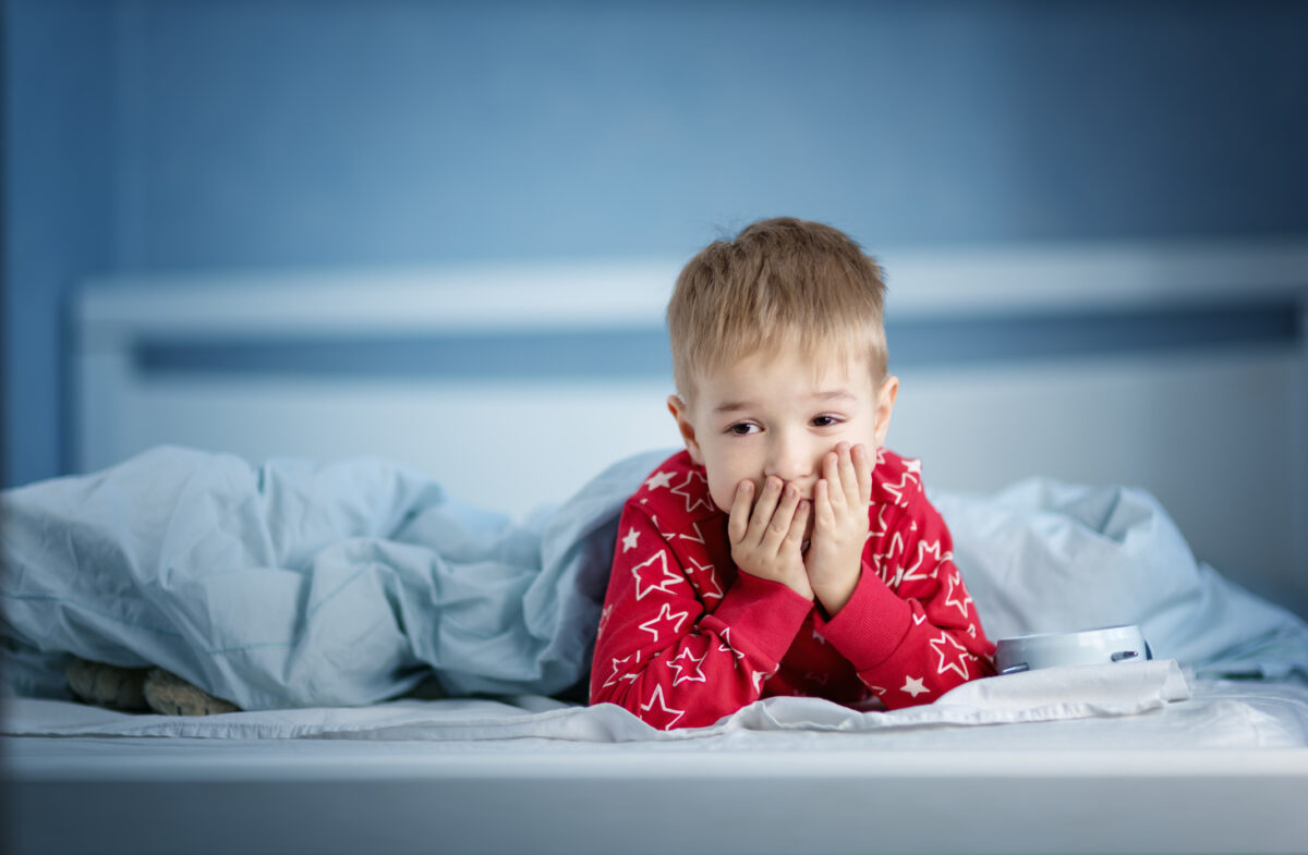 Sleepy boy lying in bed with blue beddings. Tired child in bedroom. Little kid lying awake in red pajamas with toothacheSleepy boy lying in bed with blue beddings. Tired child in bedroom sleeping. Little kid lying asleep in red pajamas with alarm clock