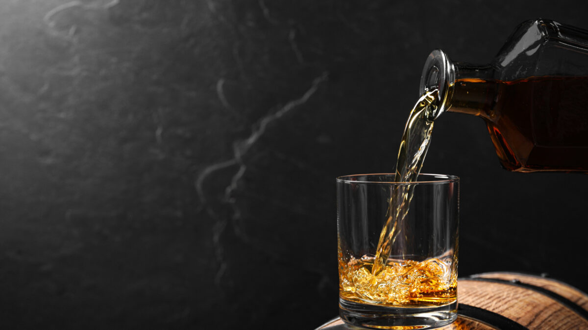 Pouring whiskey into glass from bottle on wooden barrel against grey background, space for text