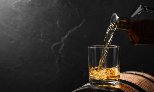 Pouring whiskey into glass from bottle on wooden barrel against grey background, space for text