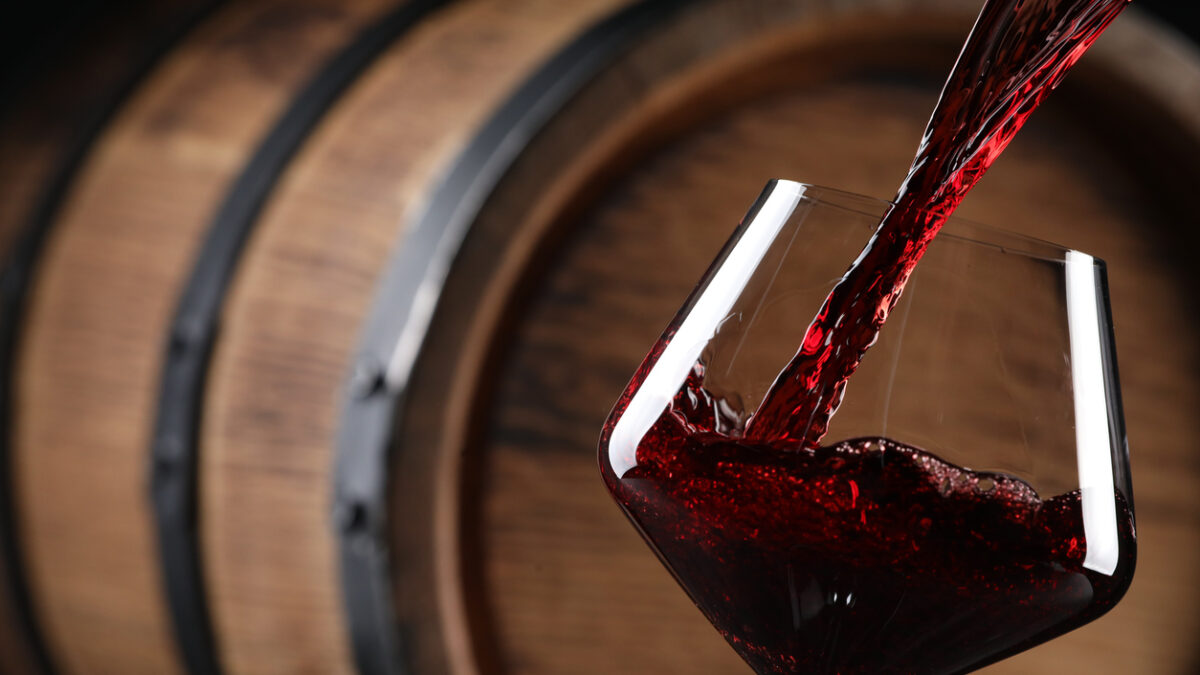 Pouring red wine into glass near wooden barrel, closeup. Space for text