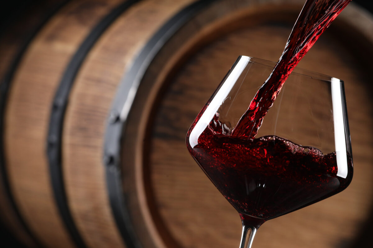 Pouring red wine into glass near wooden barrel, closeup. Space for text