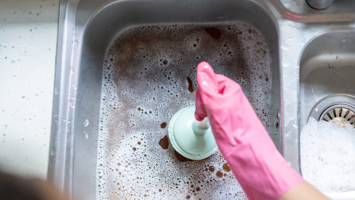 Woman Using Plunger In Blocked Kitchen Sink To Unclog Drain