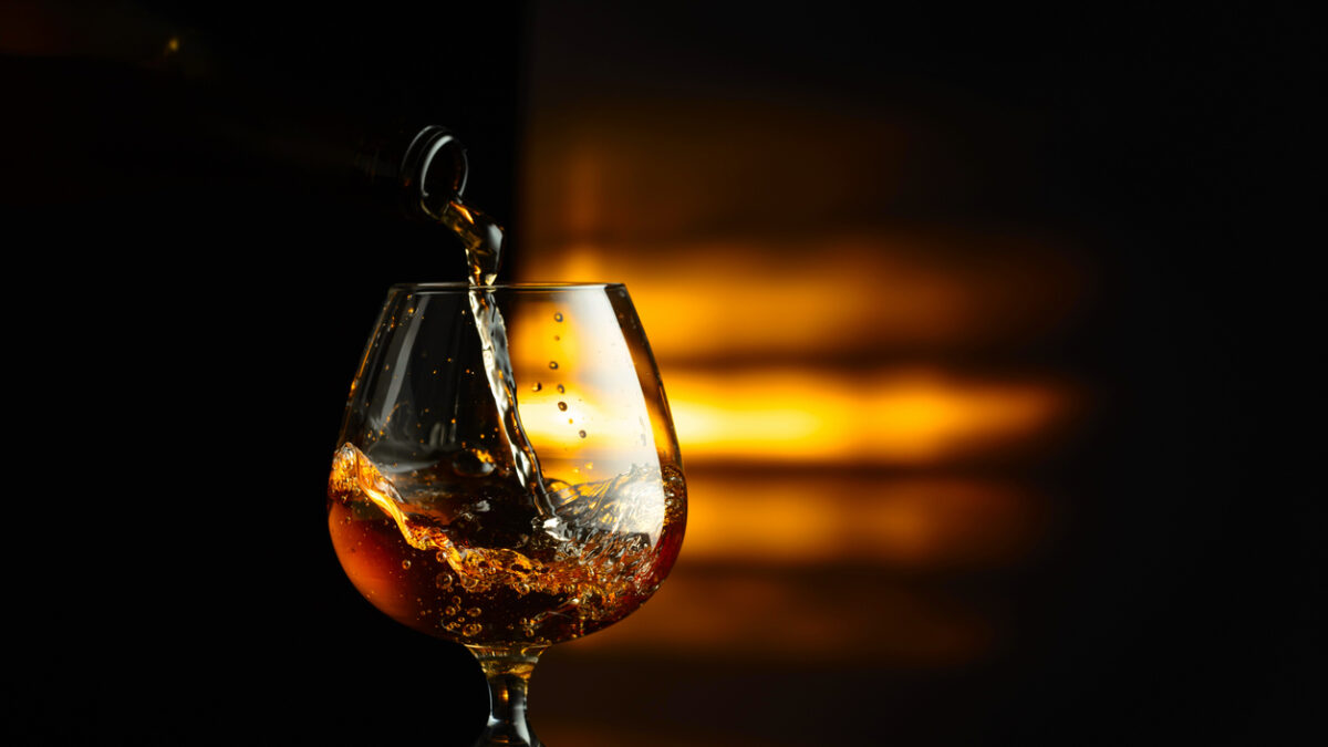 Pouring brandy from a bottle into a snifter on a black reflective background. Copy space.