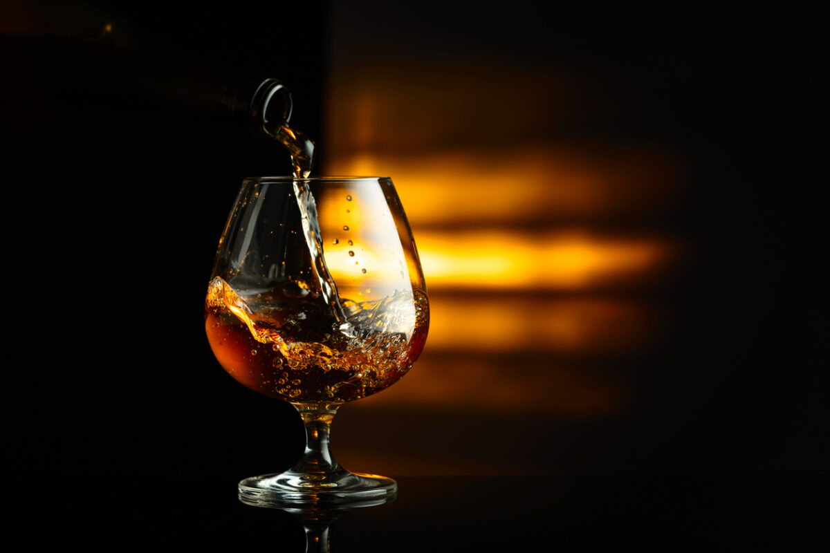 Pouring brandy from a bottle into a snifter on a black reflective background. Copy space.